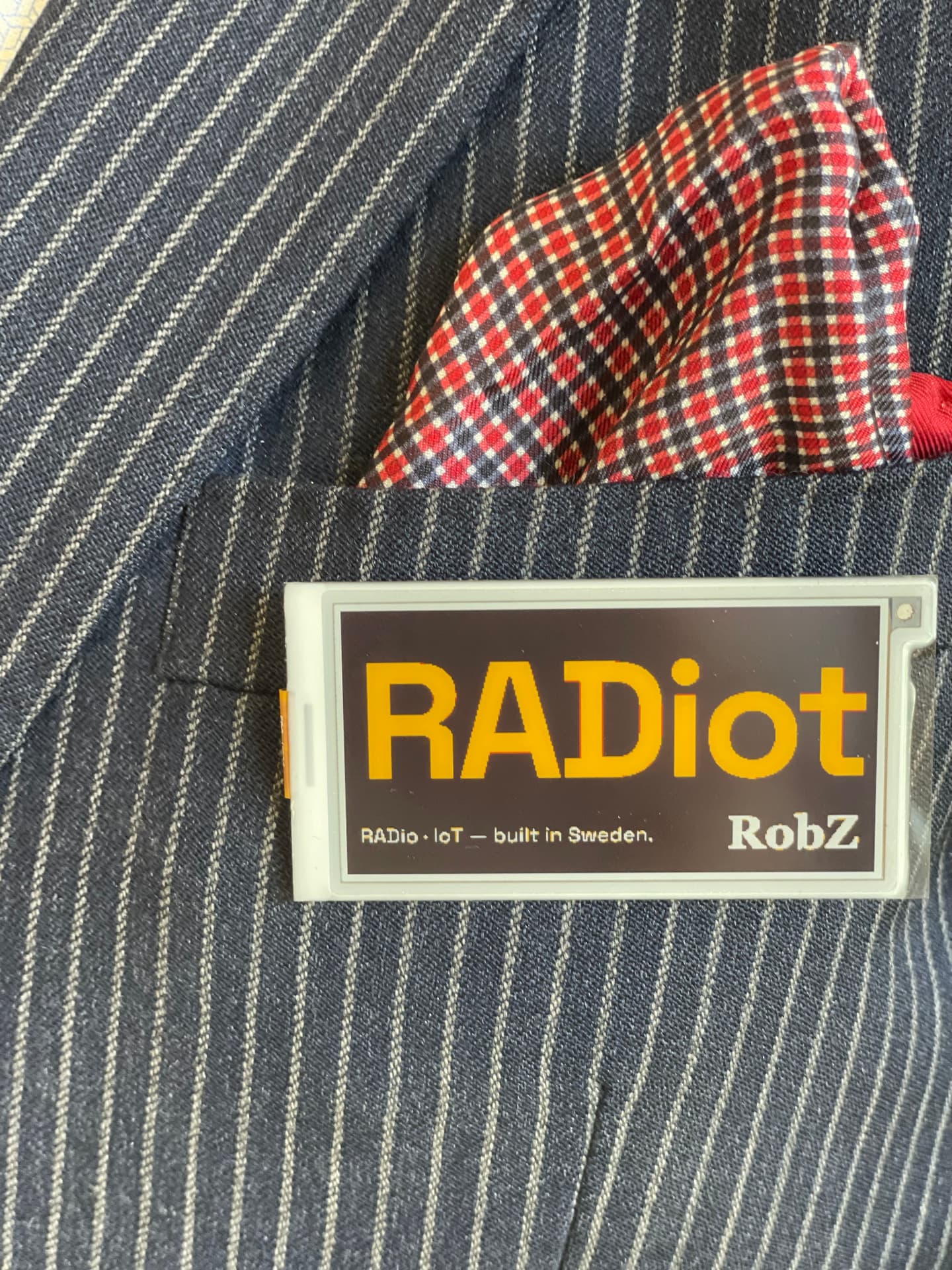 An ESTER e-paper badge worn on a pinstripe jacket pocket, showing the RADiot logo in yellow on black