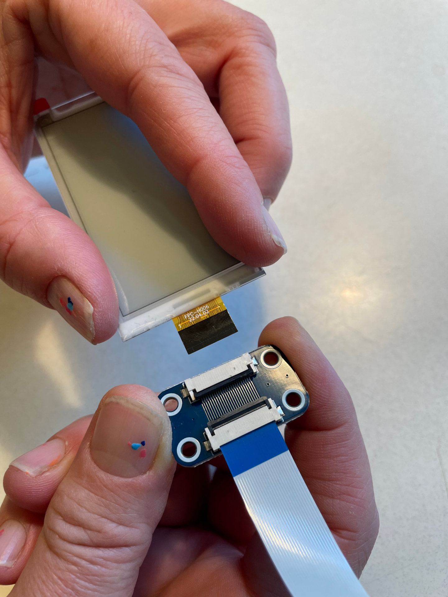 Holding the e-paper badge above the programmer board, its golden ribbon pointing at the open connector